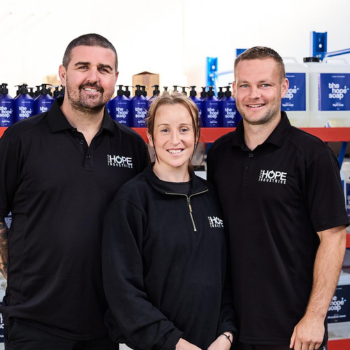 Three people in Hope Industries black polo shirts smiling together in a warehouse stocked with branded product bottles on shelving behind them.